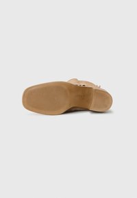 Sandale beige avec une semelle en caoutchouc texturé. Présente une forme incurvée et une sangle au talon arrière pour un maintien sécurisé. Construction robuste, design minimal.