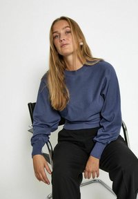 Maglione blu corto con maniche lunghe e a palloncino; texture liscia; abbinato a pantaloni neri a gamba larga. Modella seduta su una sedia moderna.