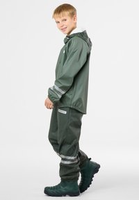 Giacca impermeabile verde con cappuccio e polsini elasticizzati, abbinata a pantaloni coordinati con strisce riflettenti e stivali in gomma testurizzati.