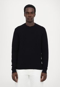 Pull en tricot noir avec un motif en losange, col rond et manches longues, associé à un pantalon blanc.