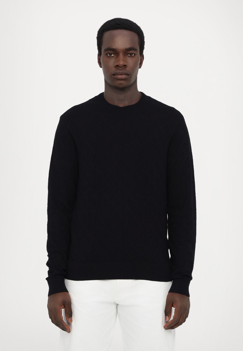 Pull en tricot noir avec un motif en losange, col rond et manches longues, associé à un pantalon blanc.