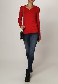 Pull rouge à col en V avec des manches longues, associé à un jean skinny bleu foncé et des bottes de cheville noires. Petite sacoche noire en bandoulière avec une chaîne.