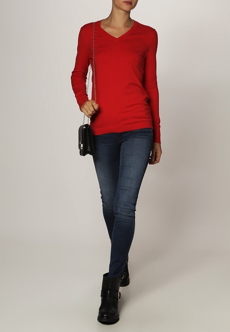 Pull rouge à col en V avec des manches longues, associé à un jean skinny bleu foncé et des bottes de cheville noires. Petite sacoche noire en bandoulière avec une chaîne.