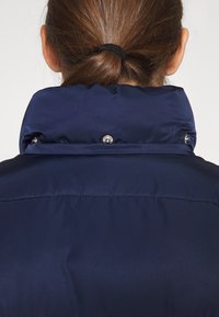 Mørkeblå dunjakke med høj krave, der har synlige trykknapper og en glat, skinnende overflade. Minimalistisk design uden mønstre.