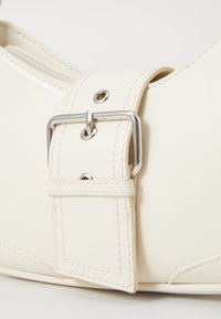 Sac à main de couleur crème avec une finition texturée, doté d'une grande boucle en métal argenté et de coutures contrastantes sur une sangle réglable.