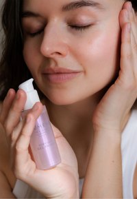 Transparent purple glass bottle of gel primer with a white pump cap, held in a hand near a face with natural skin texture.