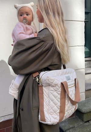 Ženska z dolgimi blond lasmi, oblečena v temen plašč, drži dojenčka v roza puloverju in klobuku z ušesi medvedka ter nosi veliko odejasto previjalno torbo z rjavimi trakovi.