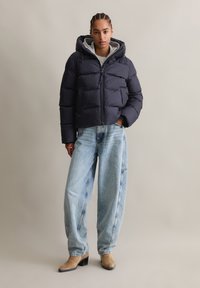Navy puffer jacket with a hood, quilted design, and front zipper. Paired with light blue wide-leg jeans and beige heeled ankle boots.