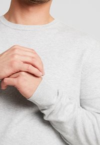 Jasnoszary sweter z dzianiny z okrągłym dekoltem, prążkowanymi mankietami i gładką teksturą. Ręce złączone z przodu, częściowo widoczne.