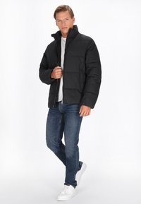 Sort puffjakke med høj krave, der har quiltede sektioner og en frontlomme. Sammen med blå jeans og hvide sneakers.