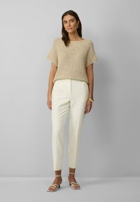 Brede beige top met een losse pasvorm, gecombineerd met crèmekleurige op maat gemaakte broeken. Het model draagt lichtgekleurde sandaaltjes met banden en een gouden armband.