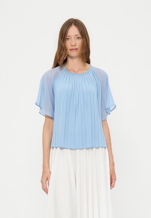 Femme aux cheveux bruns raides portant un chemisier bleu clair plissé avec des manches transparentes et une jupe blanche fluide sur un fond uni.