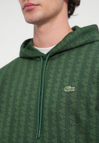 Hanorac verde cu un model geometric texturat, șnururi de culoare verde asortată și un mic logo brodat cu un crocodil pe piept.