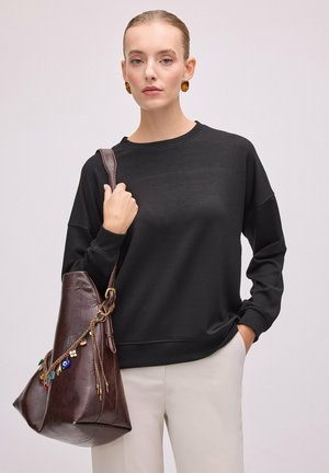Femme aux cheveux lissés en arrière, portant un chemisier noir à manches longues et un pantalon blanc, tenant un grand sac en cuir marron avec des breloques décoratives.