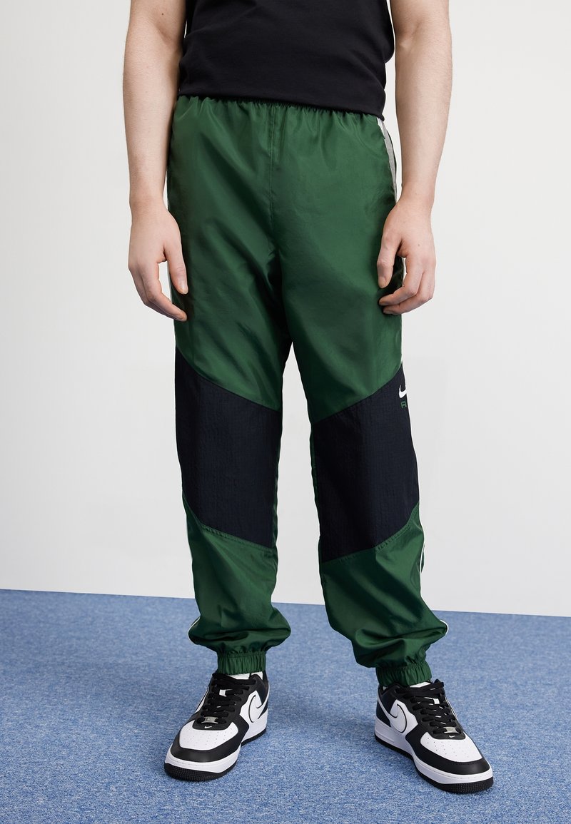 Groene en zwarte nylon trainingsbroek met elastische boorden en tailleband, met een kleurgebroken ontwerp. Draagt met zwart-witte sneakers.