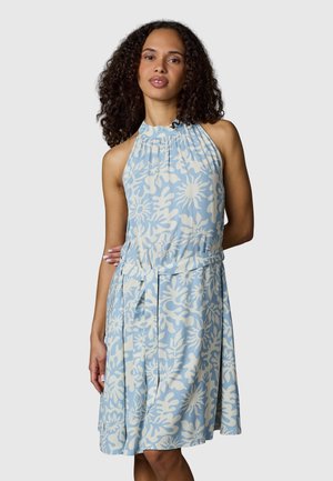 Kobieta o kręconych włosach, ubrana w jasnoniebieską sukienkę bez rękawów z białymi abstrakcyjnymi wzorami kwiatowymi i marszczonym dekoltem, stojąca z jedną ręką za plecami.