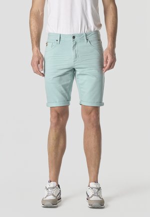 Hombre con pantalones cortos de mezclilla azul claro remangados, camiseta blanca y zapatillas blancas posando contra un fondo liso y claro.