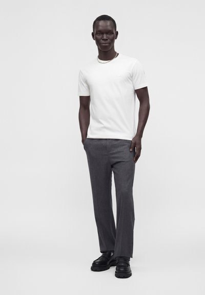 Homme portant un t-shirt blanc à manches courtes, un pantalon gris, des chaussures noires et un collier chaîne, debout les mains dans les poches.