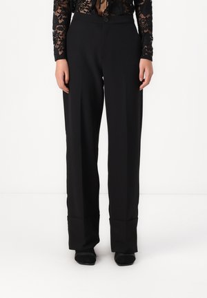 Straight-leg black trousers with front crease and cuffed hems worn with black shoes and a black lace top.