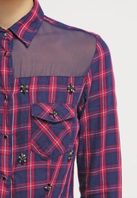 Chemise à manches longues en tartan bleu marine et rouge, dotée d'un panneau d'épaule transparent, ornée d'accents floraux noirs sur le devant et la poche.