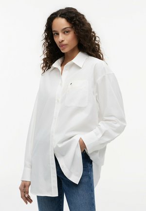 Femme aux cheveux bouclés portant une chemise blanche oversize à boutons et un jean bleu, une main dans la poche, sur fond blanc.