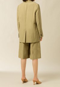 Blazer beige su misura con uno spacco singolo, abbinato a pantaloni corti larghi coordinati. Indossato con mule con tacco beige chiaro. Tessuto dalla consistenza liscia.