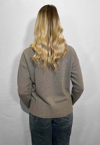 Maglione marrone a coste con scollatura rotonda e maniche lunghe, abbinato a jeans grigi a vita alta. I capelli sono ondulati e di lunghezza fino alle spalle.