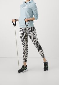 Felpa blu chiaro con logo bianco, leggings a stampa zebra in bianco e nero, scarpe da ginnastica nere con dettagli verdi, mentre tiene delle bande di resistenza.