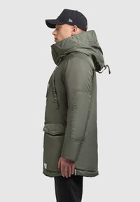 Polstret grøn parka med høj krave, stor frontlomme og justerbar hætte. Modellen har en sort kasket og mørke jeans på.