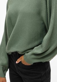 Grön ribbad tröja med långa ärmar, som har en avslappnad passform och texturerad tyg. Matchad med svarta jeans, med en hand i fickan.