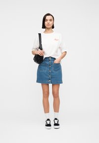 Femme portant un t-shirt blanc "Amour", une jupe en jean boutonnée bleue, un sac bandoulière noir, des chaussettes blanches et des baskets noires, debout devant un fond uni.