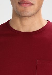 Camiseta de cuello redondo burdeos hecha de algodón suave, con un sutil detalle de bolsillo en el lado izquierdo del pecho, textura suave y costuras uniformes.