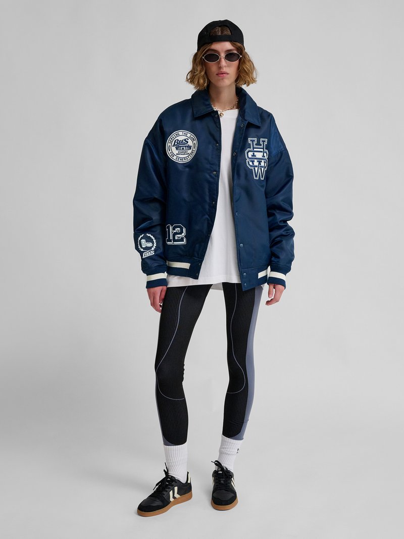 Person iført navyblå varsityjakke med mærker, hvid oversized skjorte, sorte og grå leggings, sort kasket, solbriller og sorte sneakers.