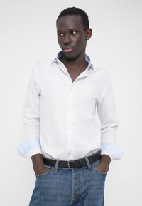 Weißes, langärmliges Hemd mit Knöpfen und hellblauen Akzenten an den Manschetten, kombiniert mit blauen Denim-Jeans und einem schwarzen Gürtel. Glatte Textur, figurbetontes Design.