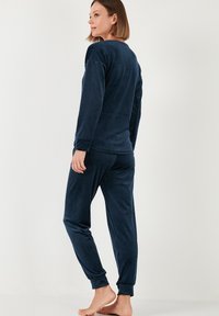 Navy blue, samettinen pyjama-setti, joka sisältää pitkähihaisen paidan ja joustavat housut. Pehmeä kangas, rento malli ja hihansuissa ja lahkeissa kuminauhakiinnitys.
