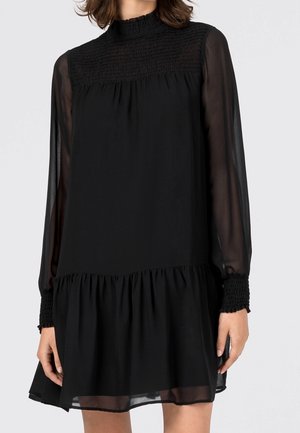 Femme portant une robe noire transparente à manches longues, avec un col haut smocké, des poignets smockés et un ourlet à volants au-dessus des genoux.