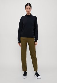 Zwarte top met hoge kraag en lange mouwen, gecombineerd met olijfgroene pantalon en zwarte sneakers met witte zolen. Eenvoudig, strak ontwerp.