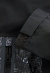 Manche de veste noire avec fermeture Velcro à côté d'une fermeture éclair noire sur un fond en tissu texturé gris et noir.
