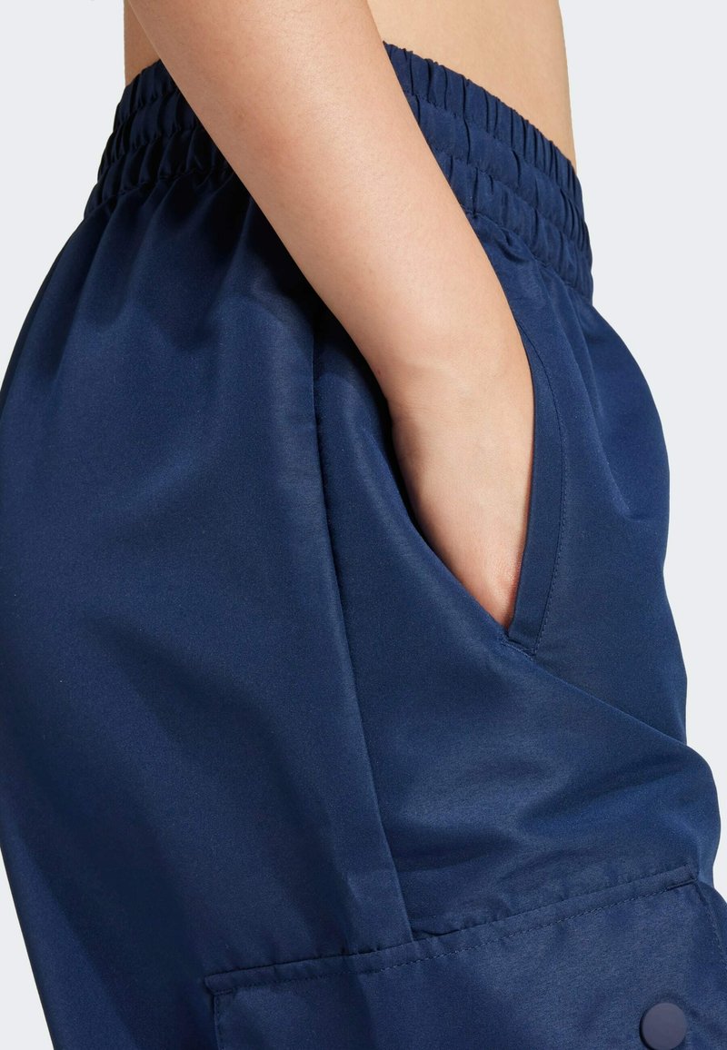 Navy blue athletic pants featuring an elastic waistband, side pocket, and smooth, shiny fabric texture. Accented with a button near the pocket.