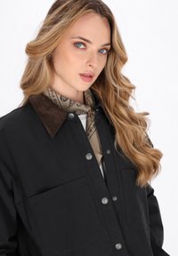 Mujer rubia con ojos azules lleva chaqueta negra con cuello marrón y bufanda de cuello de diseño beige, mirando ligeramente hacia la derecha sobre fondo blanco.
