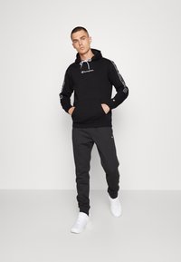 Felpa nera con logo bianco, cordino e strisce laterali, abbinata a pantaloni della tuta grigi e sneakers bianche. Design casual e aderente.