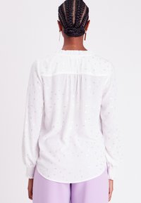 Blouse blanche avec un subtil motif de sequins argentés, manches longues avec poignets smockés, et un empiècement de dos froncé pour une touche de détail.