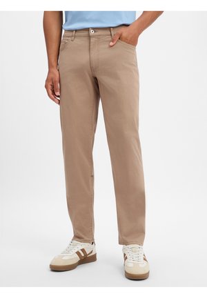 Mann trägt beige gerade geschnittene Hose und weiße Sneakers mit braunen Streifen, steht mit einer Hand in der Tasche vor einfarbigem Hintergrund.
