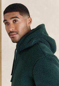 Zielona polarowa bluza z kapturem o teksturowanym wykończeniu, z regulowanym kapturem i luźnym krojem, wyeksponowana na neutralnym tle.