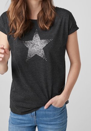 Femme portant un t-shirt gris foncé à manches courtes avec un motif étoile en sequins argentés, associé à un jean bleu clair, main dans la poche.