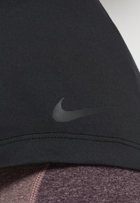 Musta urheilu paita, joka on valmistettu sileästä kankaasta ja jossa on hillitty Nike-logo tummanharmaana. Helma näyttää kontrastisen viininpunaisen, tekstuurilla varustetun kerroksen alla.