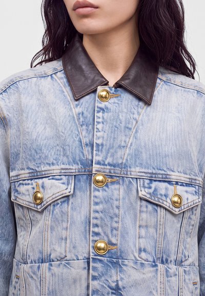 Personne portant une veste en jean bleu clair délavée à l’acide avec de gros boutons dorés et un col en cuir marron foncé, ne montrant que le cou et les épaules.