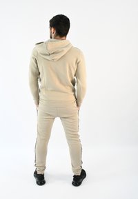 Conjunto de sudadera beige y pantalones tipo jogger fabricado en suave tejido. Los pantalones presentan rayas laterales negras. El modelo se muestra de espaldas, luciendo zapatos negros.