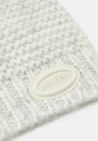 Berretto lavorato a maglia in grigio chiaro e bianco, caratterizzato da un motivo testurizzato e da una prominente etichetta ovale con il logo "GUESS". Materiale morbido e accogliente.