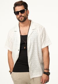 Weißes Kurzarmhemd mit eingeprägtem Blumenmuster, über einem schwarzen Tanktop getragen. Accessoires sind eine geflochtene Halskette und Armbänder.